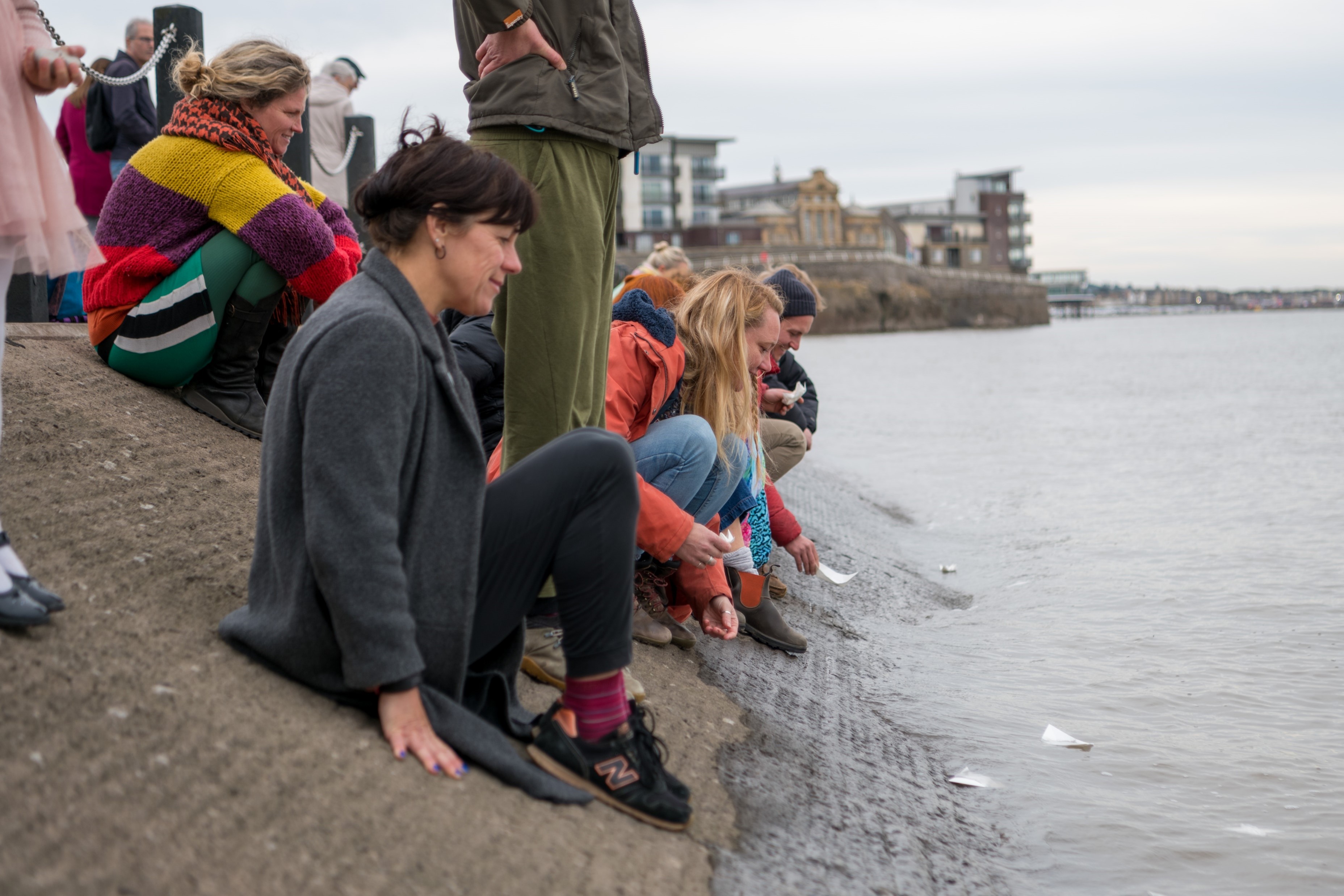 People sitting on a slope by the seafront at Weston-super-Mare watching paper offerings floating on the water.